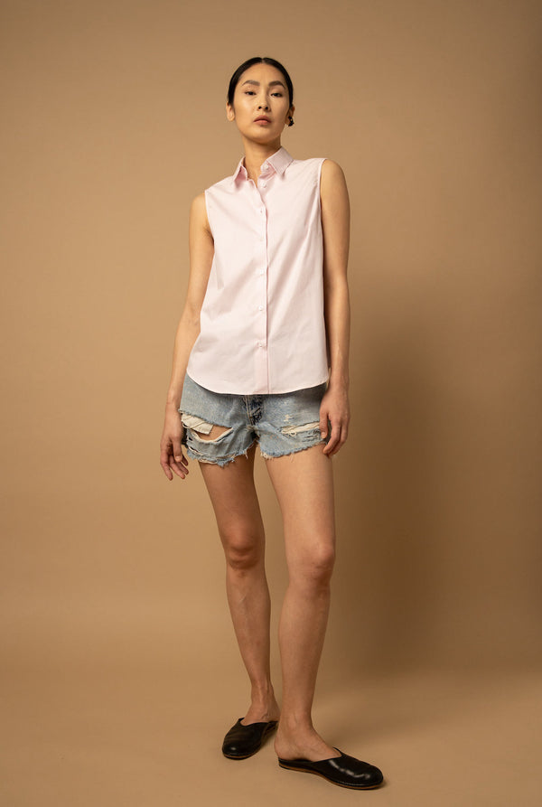 A woman wears a pink sleeveless button-down shirt paired with light-wash distressed denim shorts and a  air of mules.
