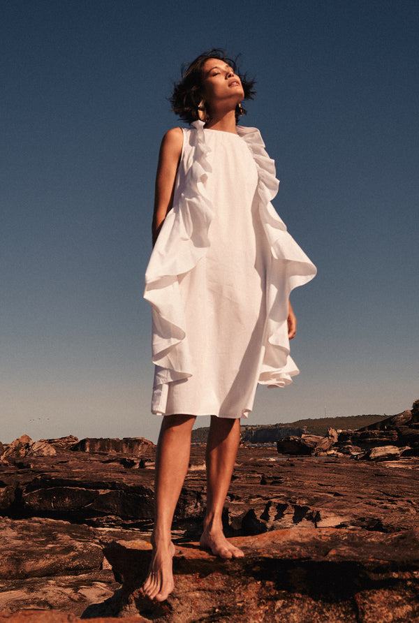  A woman wears a white ruffle dress under the sun. The ruffles run along either side of the bodice.