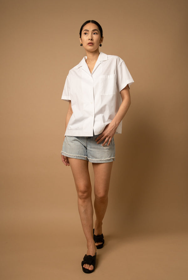  A woman wears a white poplin button-down shirt that features a classic camp collar, a breast pocket, short sleeves, and falls to the waist.