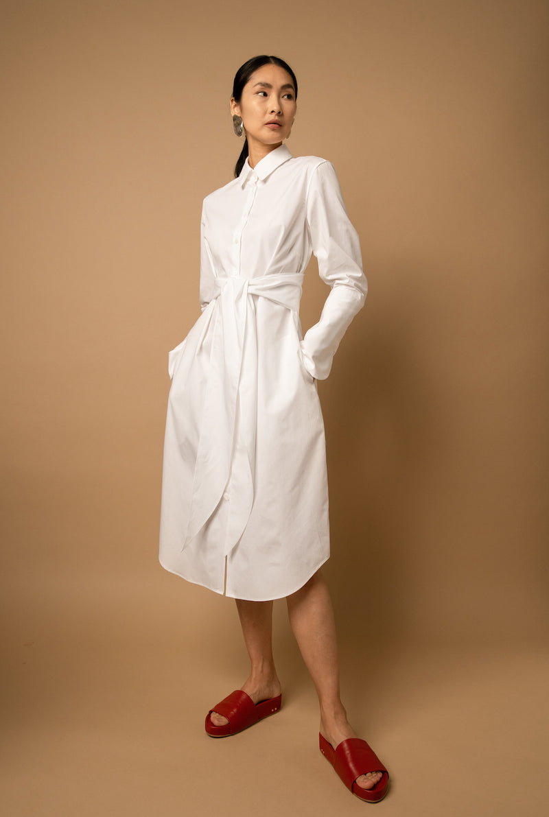 A woman wears a white poplin knee-length shirt dress that is buttoned-up all the way and has side seam pockets.