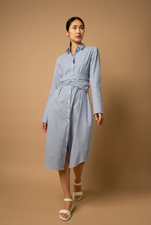 A woman wears a poplin knee-length, blue-striped shirt dress that has an A-line silhouette and long bow ties to accentuate the waist.